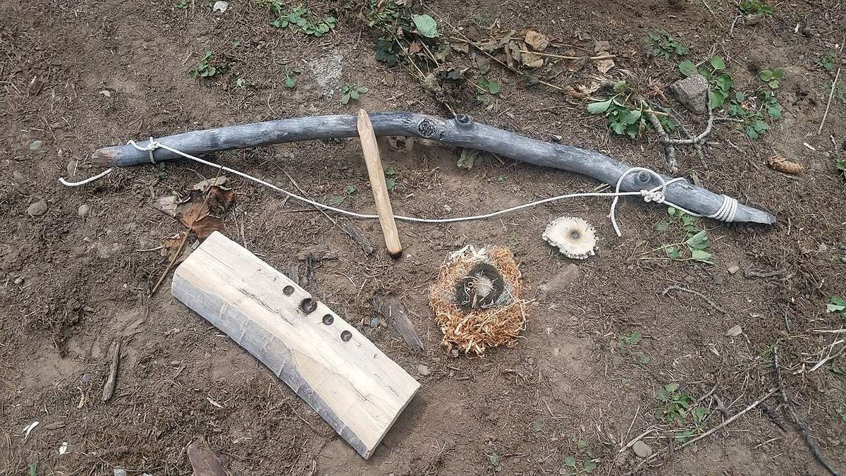 A bow, drill, notched fireboard, socket and a tinder bundle laid out on the ground