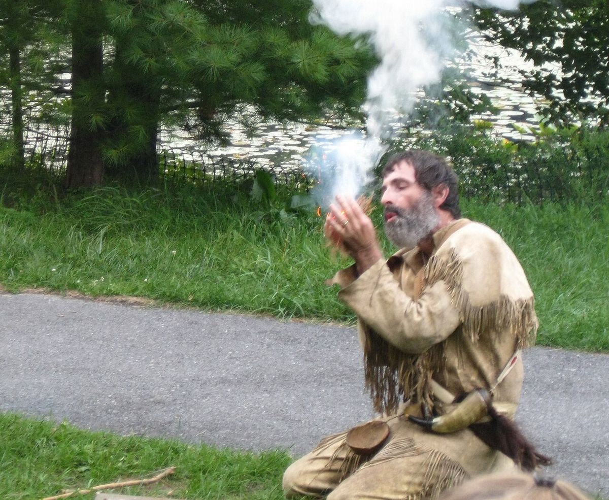 A man in buckskin blowing on smoking tinder held in his hands
