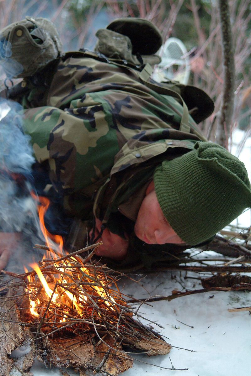 A survival student bending low to light a small fire on the snow