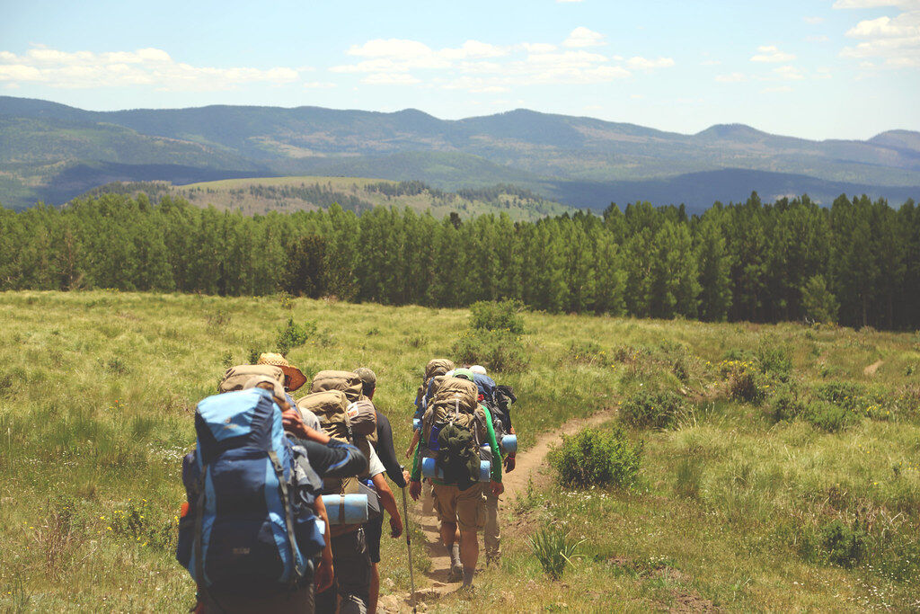 A group of hikers with big backpacks walking a dirt trail through grassland toward forested hills