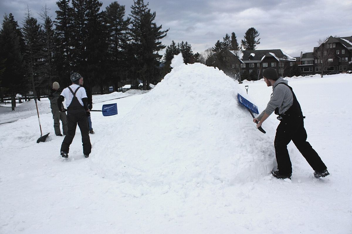 People shoveling snow into a large mound beside a parking lot