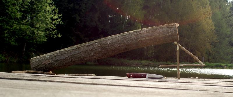 A practice figure 4 trigger of three notched sticks holding up a log on a wooden deck, with a carving knife beside it