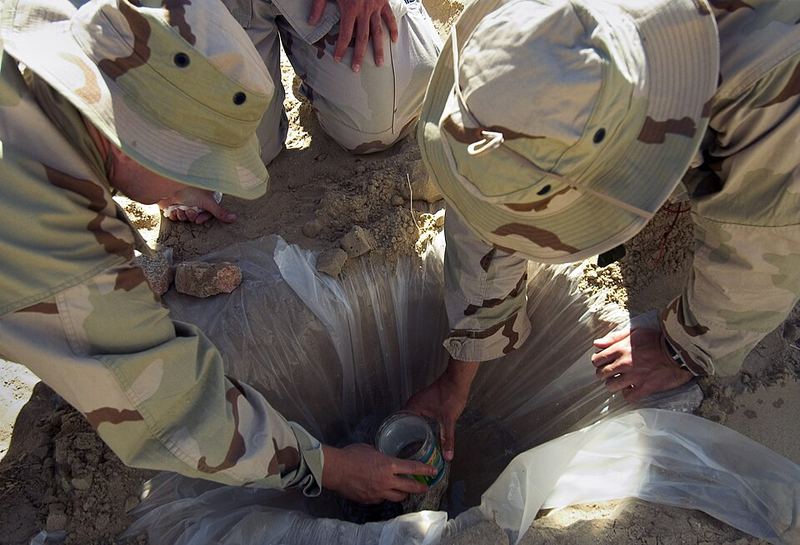 Sailors kneel around a plastic-covered hole in sand with a jar set below the center