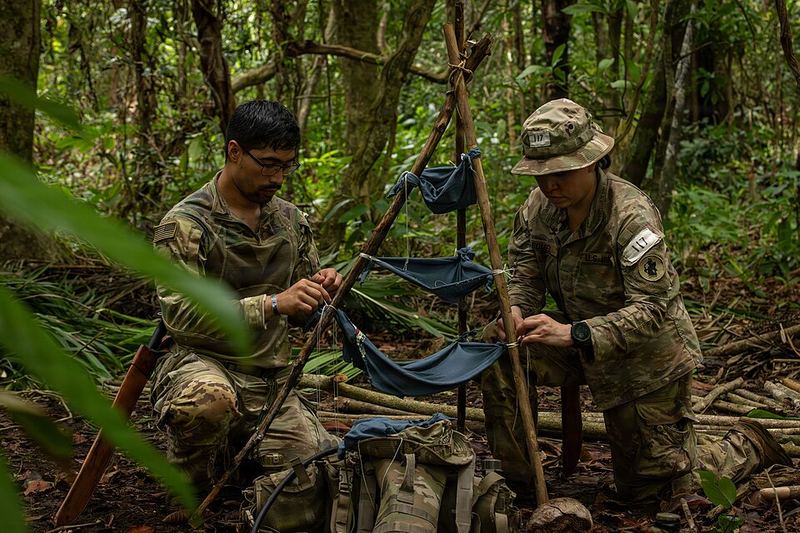 Two soldiers tie layers of cloth onto a tripod of sticks in the jungle