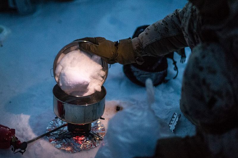 A gloved hand tips a lid of snow into a steel pot on a camp stove