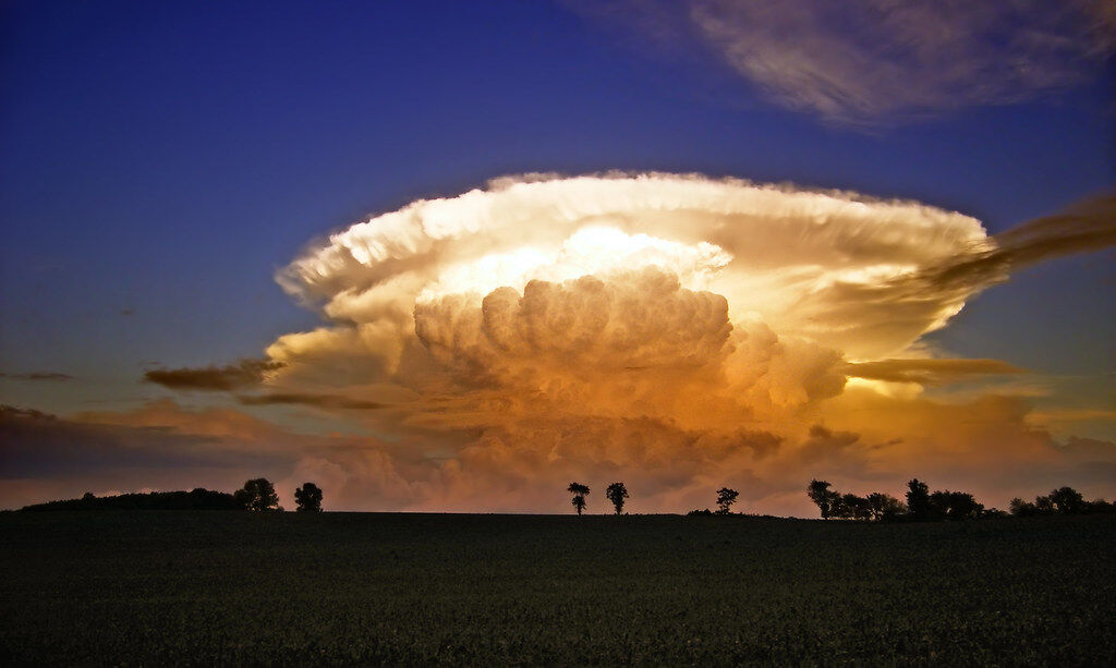 A towering thunderstorm cloud with a flat anvil top glowing at sunset over flat land